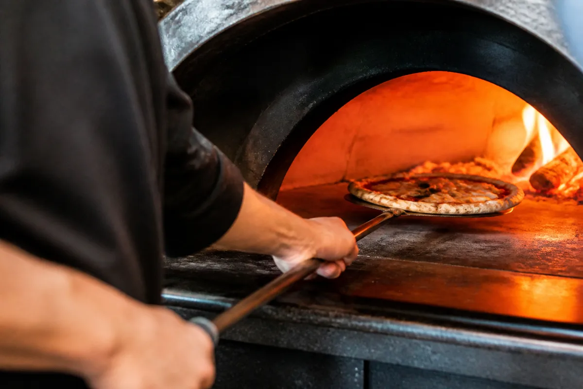 Pizzaïolo faisant pivoter une pizza à la petite pelle dans un four à bois, flamme sur le côté droit